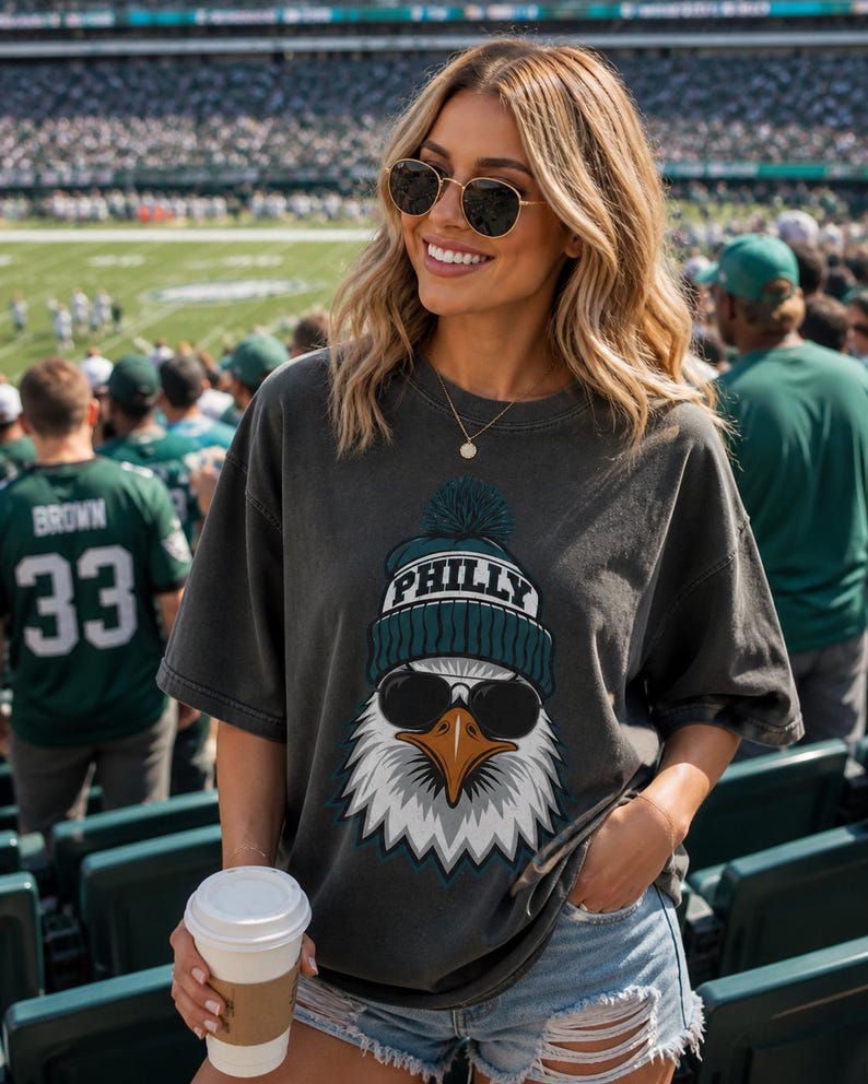 K&ouml;nnte beinhalten: Dunkelgraues T-Shirt mit einer Grafik eines Adlers, der eine Sonnenbrille und eine blaugr&uuml;ne und wei&szlig;e M&uuml;tze mit dem Wort "PHILLY" tr&auml;gt. Das Adlerdesign ist vor einem wei&szlig;en Hintergrund. Das Shirt wird mit Jeansshorts getragen.