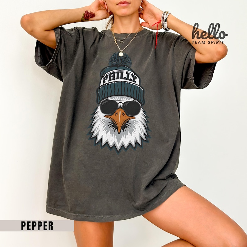 K&ouml;nnte beinhalten: Pfefferfarbenes T-Shirt mit einer Grafik eines Wei&szlig;kopfseeadlers mit Sonnenbrille und M&uuml;tze mit der Aufschrift "PHILLY". Der Adler hat einen wei&szlig;-grauen gefiederten Kopf und einen braunen Schnabel. Das Shirt hat kurze &Auml;rmel und eine lockere Passform.