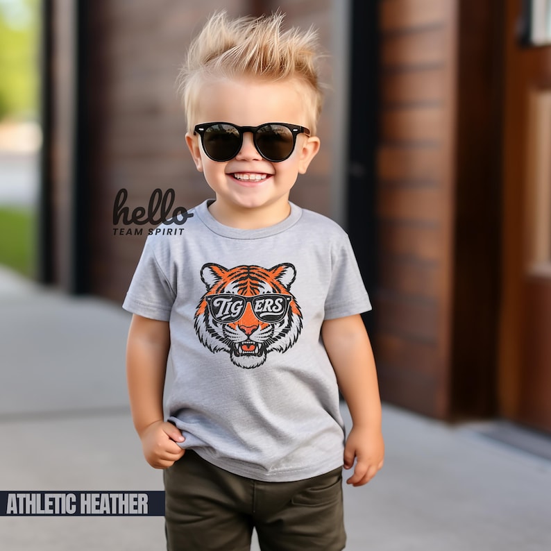 Peut inclure: Un jeune enfant porte un t-shirt gris chin&eacute; avec un motif de tigre et le mot "Tigers" sur des lunettes de soleil noires. Le t-shirt porte &eacute;galement le texte "hello team spirit". L'enfant porte &eacute;galement des lunettes de soleil noires et un pantalon kaki.