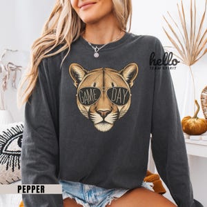 May include: Dark gray sweatshirt featuring a cougar wearing sunglasses with the words "GAME DAY." The sweatshirt has long sleeves and a crew neck. The word "hello" and "TEAM SPIRIT" are printed on the upper right side. The word "PEPPER" is printed on the bottom left.