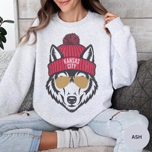 Peut inclure: Un sweat-shirt gris avec un loup portant un bonnet rouge et blanc avec le texte "Kansas City" dessus. Le loup porte des lunettes de soleil.