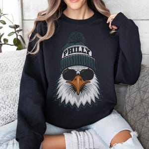 K&ouml;nnte beinhalten: Marineblaues Sweatshirt mit einer Grafik eines Adlers, der eine Sonnenbrille und eine gr&uuml;n-wei&szlig;e Strickm&uuml;tze mit dem Wort "PHILLY" tr&auml;gt. Der Adler hat wei&szlig;e Federn und einen braunen Schnabel. Rundhalsausschnitt.