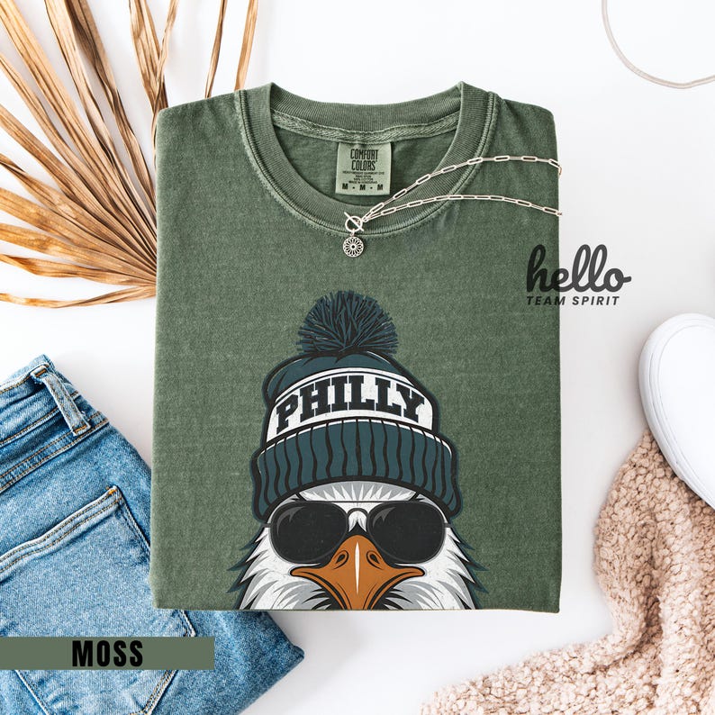 K&ouml;nnte beinhalten: Moosgr&uuml;nes T-Shirt mit einer Grafik eines Adlers, der eine M&uuml;tze mit "PHILLY" und Sonnenbrille tr&auml;gt. Das Shirt ist gefaltet mit einer silbernen Halskette und einer Jeans. Die Worte "hello TEAM SPIRIT" sind auf der rechten Seite aufgedruckt.