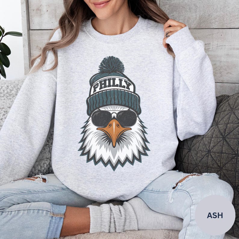 K&ouml;nnte beinhalten: Ein hellgraues Sweatshirt mit einer Grafik eines Adlers, der eine t&uuml;rkisfarbene M&uuml;tze mit der Aufschrift "PHILLY" und eine Sonnenbrille tr&auml;gt. Der Adler hat ein wei&szlig;es und graues Federdesign mit einem orangefarbenen Schnabel. Das Sweatshirt ist ein Rundhals-Stil.