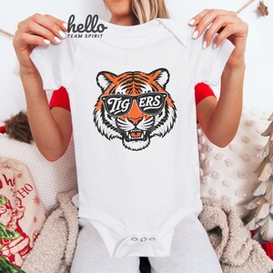 Peut inclure: Body b&eacute;b&eacute; blanc avec un motif de tigre portant des lunettes de soleil. Le tigre est orange, noir et blanc, avec le mot "TIGERS" sur les lunettes. Le body a des manches courtes et des boutons-pression.