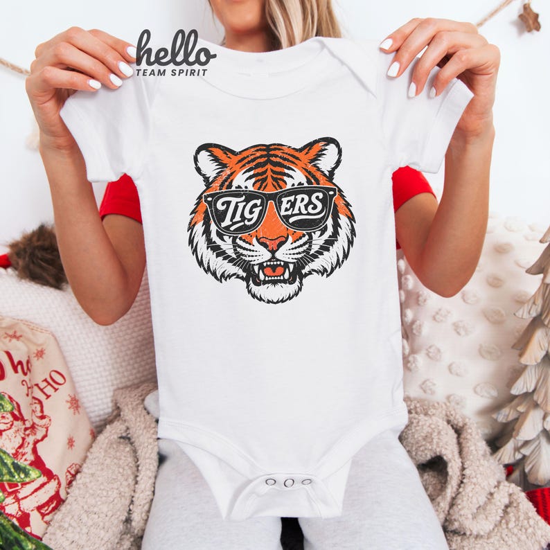 Peut inclure: Body b&eacute;b&eacute; blanc avec un motif de tigre portant des lunettes de soleil. Le tigre est orange, noir et blanc, avec le mot "TIGERS" sur les lunettes. Le body a des manches courtes et des boutons-pression.