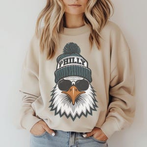 K&ouml;nnte beinhalten: Beiges Sweatshirt mit einem Aufdruck eines Adlers, der eine tealfarbene M&uuml;tze mit dem Wort "PHILLY" und Sonnenbrille tr&auml;gt. Der Adler hat ein wei&szlig;es und graues Federdesign mit einem orangefarbenen Schnabel. Das Sweatshirt hat lange &Auml;rmel.