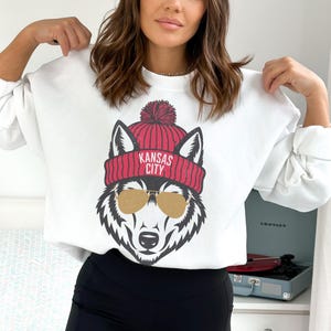 Peut inclure: Sweat-shirt blanc avec un motif de loup portant un bonnet rouge avec le texte "KANSAS CITY" et des lunettes de soleil.