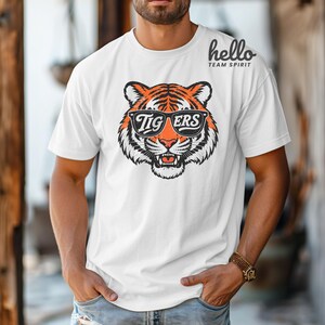 Peut inclure: T-shirt blanc avec un motif de t&ecirc;te de tigre portant des lunettes de soleil avec l'inscription "Tigers". Le tigre a des rayures orange et noires. Le t-shirt porte &eacute;galement le texte "hello TEAM SPIRIT" dans le coin sup&eacute;rieur droit.