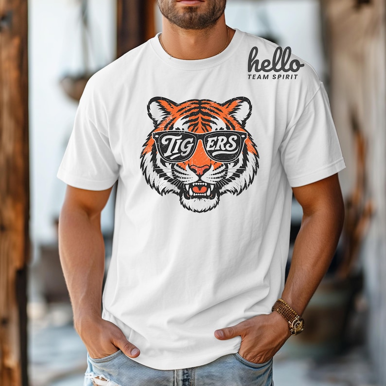 Peut inclure: T-shirt blanc avec un motif de t&ecirc;te de tigre portant des lunettes de soleil avec l'inscription "Tigers". Le tigre a des rayures orange et noires. Le t-shirt porte &eacute;galement le texte "hello TEAM SPIRIT" dans le coin sup&eacute;rieur droit.