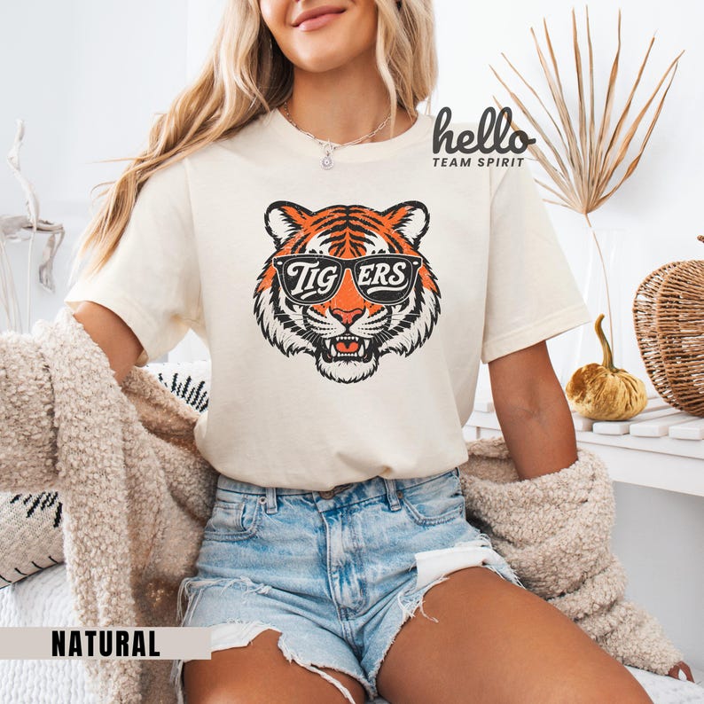 Peut inclure: T-shirt cr&egrave;me avec une t&ecirc;te de tigre portant des lunettes de soleil avec le mot "TIGERS". Le tigre a des rayures orange et noires. Le t-shirt est &eacute;tiquet&eacute; "NATURAL" en bas.