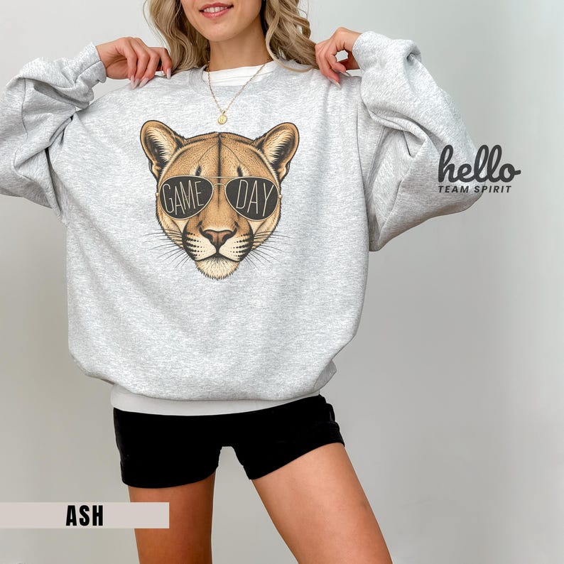 May include: Ash-colored sweatshirt featuring a cougar face wearing sunglasses with "GAME DAY" text. The sweatshirt has long sleeves and a crew neck. The words "hello TEAM SPIRIT" are printed on the right sleeve.