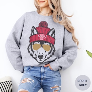 Peut inclure: Un sweat-shirt gris avec un loup portant un bonnet rouge avec le texte "Kansas City" dessus. Le loup porte des lunettes de soleil.