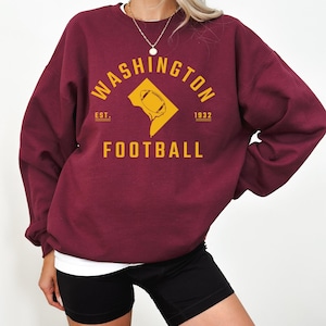 May include: Burgundy sweatshirt with a gold graphic of the Washington D.C. outline with a football inside. The text "WASHINGTON" is in an arc above the outline and "EST. 1932" is below the outline. The text "FOOTBALL" is below the graphic.