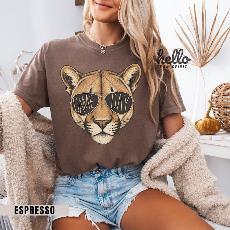 May include: Espresso-colored t-shirt featuring a cougar wearing sunglasses with the words "GAME DAY" printed on the lenses. The shirt has a crew neck and short sleeves. The text "hello TEAM SPIRIT" is printed on the upper right side.
