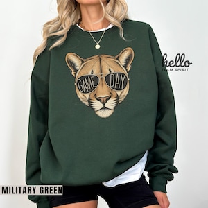 May include: Military green sweatshirt featuring a cougar head wearing sunglasses with the words "GAME DAY". The sweatshirt has a crew neck and long sleeves. The text "MILITARY GREEN" is at the bottom of the image.