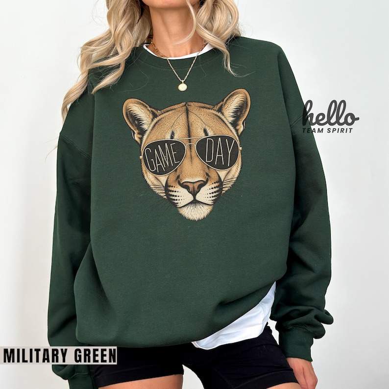 May include: Military green sweatshirt featuring a cougar head wearing sunglasses with the words "GAME DAY". The sweatshirt has a crew neck and long sleeves. The text "MILITARY GREEN" is at the bottom of the image.