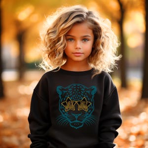 May include: A young girl wearing a black sweatshirt with a blue and black leopard print graphic. The leopard is wearing gold star-shaped sunglasses with the text "Jax Jags" written on them.