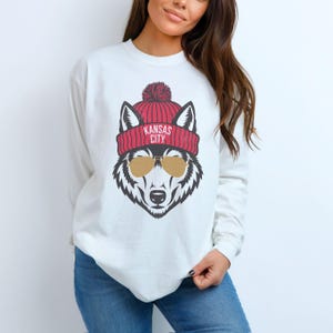 May include: White sweatshirt with a graphic of a wolf wearing a red and white beanie with the text "Kansas City" on it. The wolf is also wearing sunglasses.