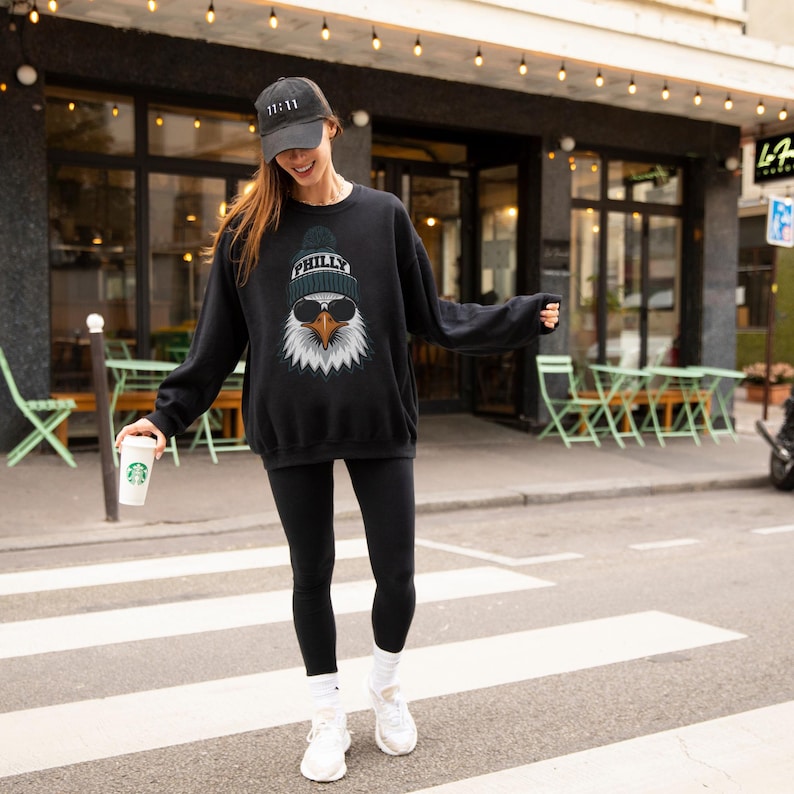 K&ouml;nnte beinhalten: Schwarzes Sweatshirt mit einem Adler-Motiv, das eine Sonnenbrille und eine M&uuml;tze mit dem Wort "PHILLY" tr&auml;gt. Die Person tr&auml;gt auch eine schwarze Baseballkappe mit dem Text "11:11". Die Person tr&auml;gt schwarze Leggings und wei&szlig;e Turnschuhe.