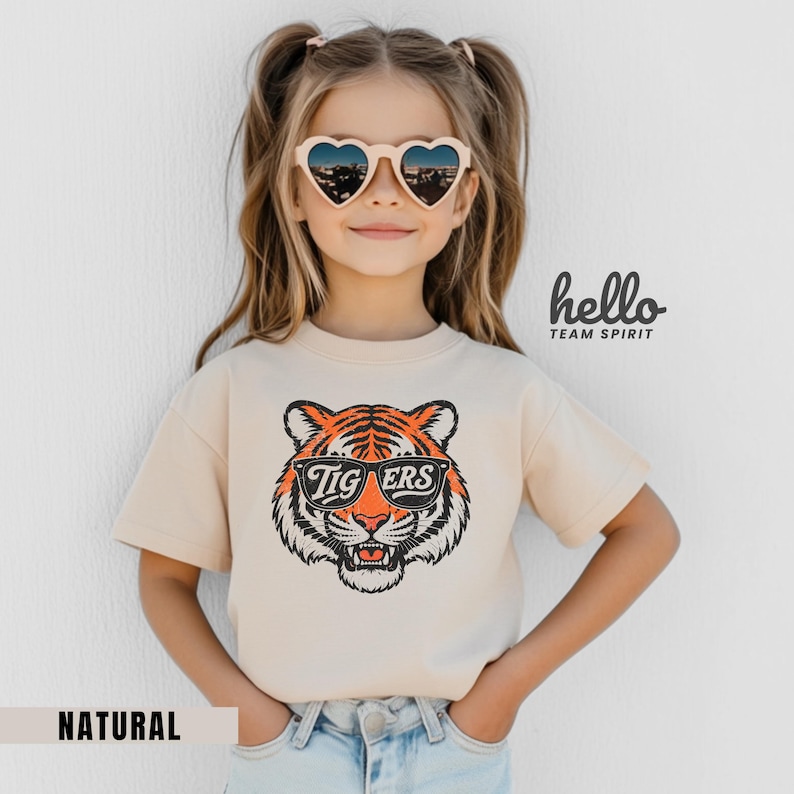 Peut inclure: T-shirt beige &agrave; manches courtes avec un motif de tigre portant des lunettes de soleil et le mot "Tigers". Le mot "Natural" est en bas. Les mots "hello team spirit" sont sur le c&ocirc;t&eacute; droit.
