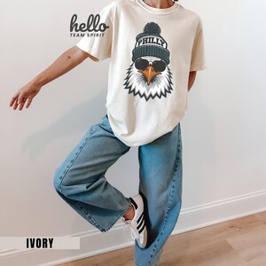 K&ouml;nnte beinhalten: Elfenbeinfarbenes T-Shirt mit einer Grafik eines Adlers mit M&uuml;tze und Sonnenbrille. Auf der M&uuml;tze steht "PHILLY". Auf dem T-Shirt steht links oben "hello TEAM SPIRIT". Das Wort "IVORY" ist unten aufgedruckt.