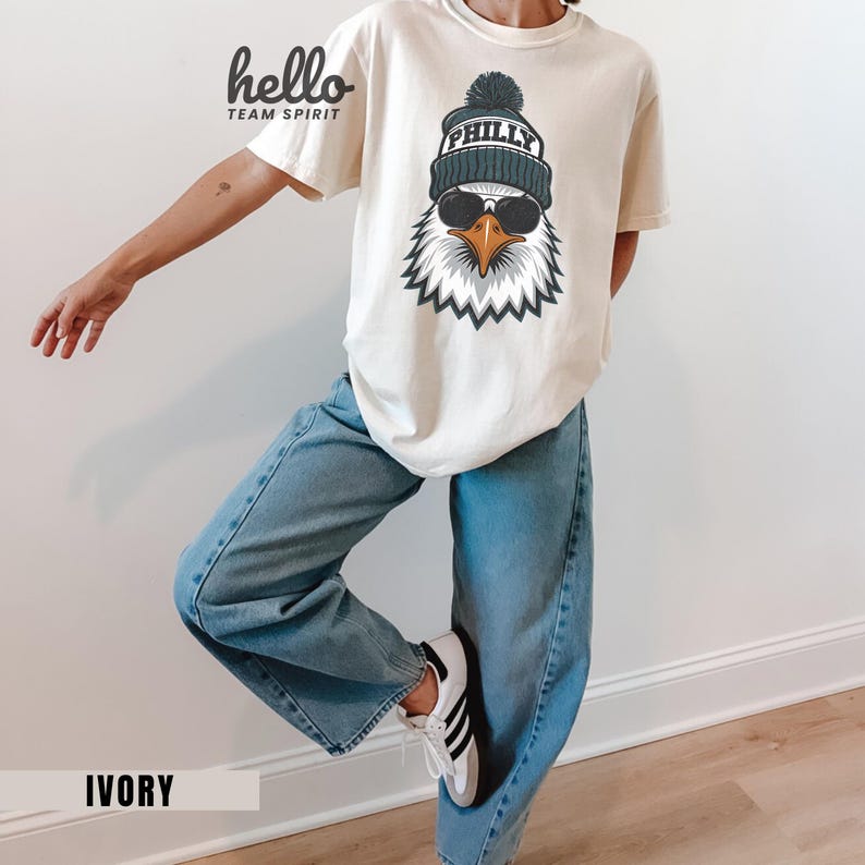 K&ouml;nnte beinhalten: Elfenbeinfarbenes T-Shirt mit einer Grafik eines Adlers mit M&uuml;tze und Sonnenbrille. Auf der M&uuml;tze steht "PHILLY". Auf dem T-Shirt steht links oben "hello TEAM SPIRIT". Das Wort "IVORY" ist unten aufgedruckt.