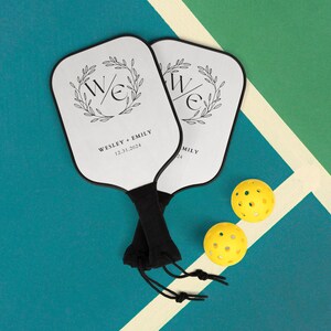 May include: Two white paddleball rackets with black handles and black trim. The rackets have a black and white design with the letters "W" and "E" in a wreath. The text "WESLEY + EMILY 12.31.2024" is printed on one racket. Two yellow paddleballs are on a blue and green surface.