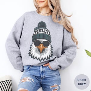 K&ouml;nnte beinhalten: Sportgraues Sweatshirt mit einem Adler-Grafik, der eine Sonnenbrille und eine tealfarbene M&uuml;tze mit der Aufschrift "PHILLY" tr&auml;gt. Der Adler hat wei&szlig;e und graue Federn und einen braunen Schnabel. Das Sweatshirt wird mit einer blauen Jeans getragen.