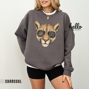 May include: Charcoal gray sweatshirt featuring a cougar wearing sunglasses with the text "GAME DAY". The sweatshirt has a crew neck and long sleeves. The words "hello TEAM SPIRIT" are printed on the upper right side.