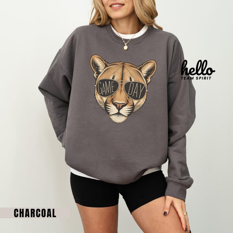 May include: Charcoal gray sweatshirt featuring a cougar wearing sunglasses with the text "GAME DAY". The sweatshirt has a crew neck and long sleeves. The words "hello TEAM SPIRIT" are printed on the upper right side.