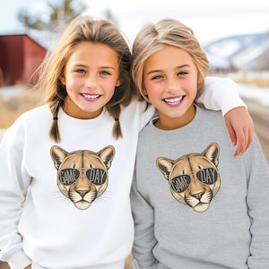 May include: Two children wearing sweatshirts. One sweatshirt is white, the other is gray. Both sweatshirts feature a cougar head wearing sunglasses with the text "GAME DAY". The children are smiling.