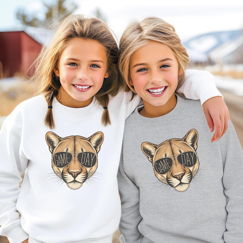 May include: Two children wearing sweatshirts. One sweatshirt is white, the other is gray. Both sweatshirts feature a cougar head wearing sunglasses with the text "GAME DAY". The children are smiling.