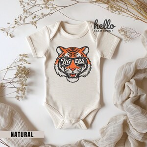 Peut inclure: Un body de b&eacute;b&eacute; cr&egrave;me avec un motif de tigre portant des lunettes de soleil avec le mot "TIGERS" sur les verres. Le texte "hello TEAM SPIRIT" est imprim&eacute; en haut &agrave; droite. Le mot "NATURAL" est imprim&eacute; en bas &agrave; gauche.
