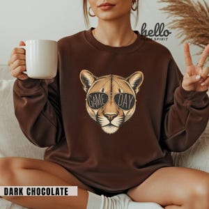 May include: Dark chocolate brown sweatshirt featuring a cougar head wearing sunglasses with the words "GAME DAY". The sweatshirt has a crew neck and long sleeves. The text "DARK CHOCOLATE" is at the bottom of the image.