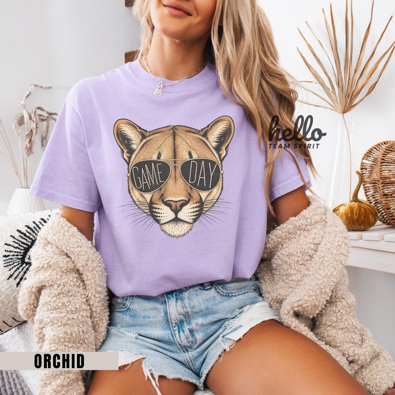 May include: A light purple t-shirt featuring a cougar head wearing sunglasses with the words "GAME DAY" printed on the lenses. The shirt is labeled "ORCHID" at the bottom. The design is centered on the front of the shirt.