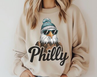 May include: A beige sweatshirt with a graphic of an eagle wearing a blue and white winter hat and sunglasses. The text "Philly" is printed in black below the eagle.