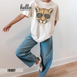 May include: Ivory-colored t-shirt featuring a cougar wearing sunglasses with the text "GAME DAY." The shirt also has the text "hello TEAM SPIRIT" in black. The person is wearing light blue jeans and white sneakers with black stripes.