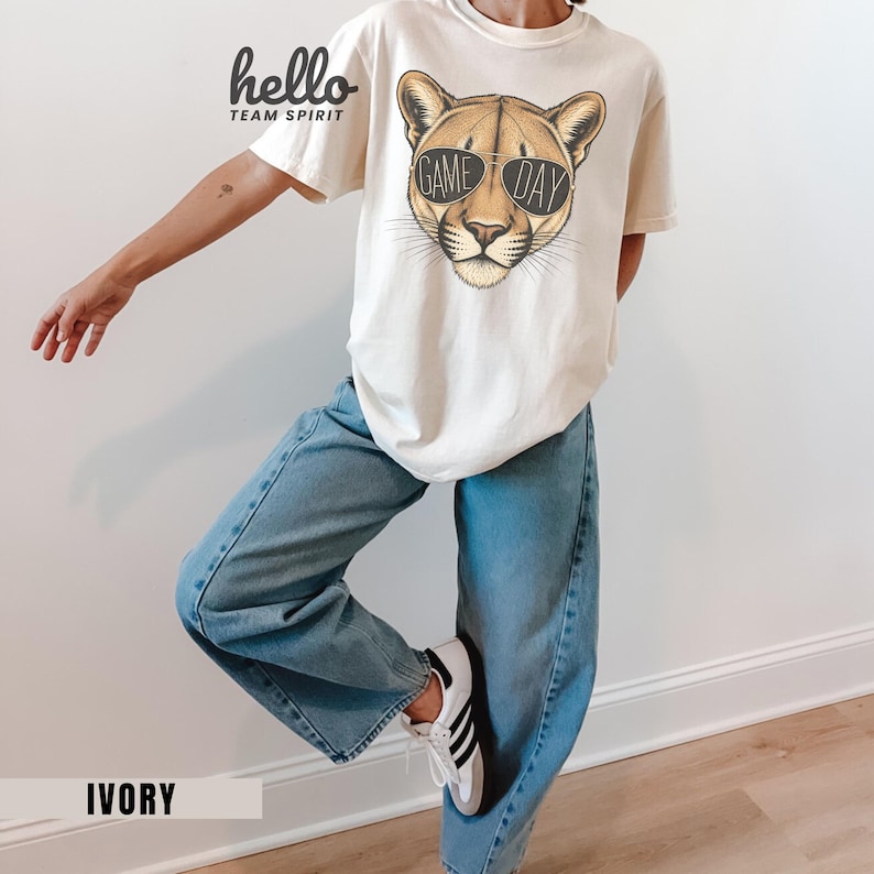 May include: Ivory-colored t-shirt featuring a cougar wearing sunglasses with the text "GAME DAY." The shirt also has the text "hello TEAM SPIRIT" in black. The person is wearing light blue jeans and white sneakers with black stripes.