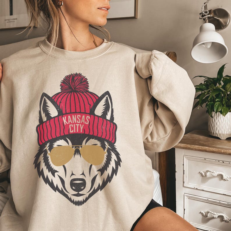 Peut inclure: Un sweat-shirt beige avec un motif de loup portant un bonnet rouge et blanc en tricot avec le texte "Kansas City" sur le bonnet. Le loup porte des lunettes de soleil &agrave; monture dor&eacute;e.