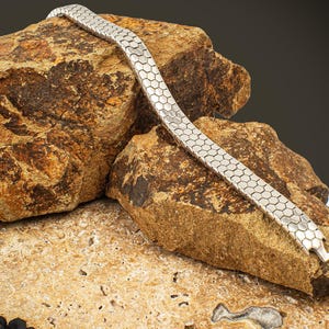 Puede incluir: Una pulsera plateada con un diseño de panal descansa sobre una superficie rocosa texturizada. El diseño de la pulsera presenta una serie de formas hexagonales interconectadas, creando un accesorio flexible y llamativo. Una joya elegante.