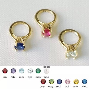 May include: Three gold-toned ring charms featuring prong-set round gemstones in blue, pink, and clear. Below, a color-coded chart displays twelve birthstone options labeled with months from January to December, including pearl for June.