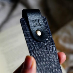 May include: A close-up of a black leather bookmark with a textured, crocodile-like pattern. The bookmark has a loop at the top with the initials "MK" embossed. A silver-colored rivet secures the loop. The bookmark is held in front of a book.