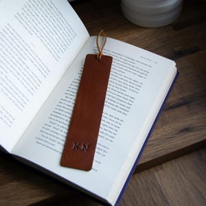 May include: A brown leather bookmark with the initials "KN" embossed on it. The bookmark is lying on top of an open book.