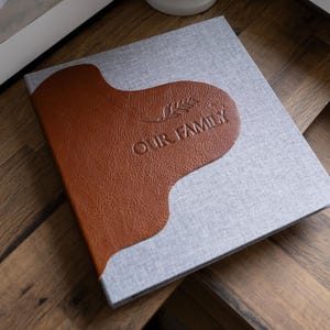 Peut inclure: Un album photo gris et marron avec les mots "Our Family" en relief sur la couverture.