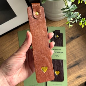 May include: A brown leather bookmark with a gold heart and a snap closure. The bookmark is rectangular and has a textured surface. The bookmark is approximately 7 inches long.