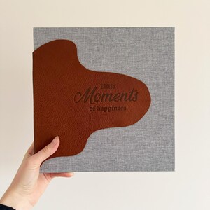 May include: A square photo album with a gray fabric cover and a brown leather accent. The words "Little Moments of happiness" are embossed on the cover in a script font. The album is held by a person.