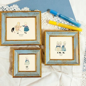 May include: Three framed illustrations with light blue and gold distressed frames. The illustrations depict children in vintage clothing. Two markers are visible in the background. The text in the illustrations is in a foreign language.