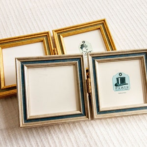 Könnte beinhalten: Zwei quadratische, klappbare Bilderrahmen. Ein Rahmen ist goldfarben mit grünen Akzenten, der andere silberfarben mit blauem Rand. Die Rahmen sind leer, mit weißer Rückseite. Ein Rahmen hat ein Etikett mit "PARIS" und einer Hut-Illustration.