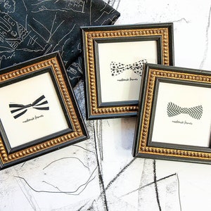 Könnte beinhalten: Drei gerahmte Fliegen-Drucke mit schwarzen und goldenen, verzierten Rahmen. Jeder Druck zeigt ein anderes Fliegen-Design in Schwarz und Weiß. Der Text "mackenzie frames" ist unter jedem Fliegen-Design gedruckt.